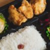 Un bento ou boite à repas japonais dans laquelle on peut voir du riz blanc cuit, des légumes marinés et du poulet pané et frit avec un peu de sauce