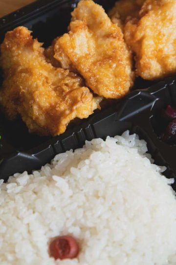 Un bento ou boite à repas japonais dans laquelle on peut voir du riz blanc cuit, des légumes marinés et du poulet pané et frit avec un peu de sauce