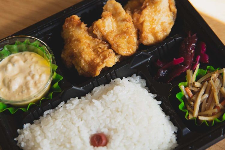 Un bento ou boite à repas japonais dans laquelle on peut voir du riz blanc cuit, des légumes marinés et du poulet pané et frit avec un peu de sauce