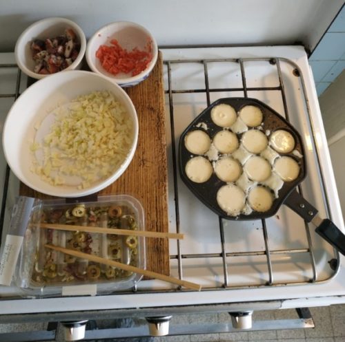 une gazinière sur laquelle est posée des bols avec tous les ingrédients sur la gauche pour contenir les ingrédients pour faire des takoyaki. À droite, on voit une poêle à takoyaki, dont les trous sont remplis de pâte à takoyaki. Les takoyaki sont en train de cuire.